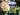 Person outdoors wearing sunglasses, headphones, a white cap that reads DONT DIE. HAVE FUN. SKI FAST., and a green jacket, with blurred trees in the background.