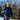 A man in a neon cap, gloves, and denim jacket stands on snow with trees behind him, holding ski poles and wearing Julbo-usa Explorer 2.0 glacier sunglasses with high-protection lenses.