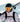 A skier wearing a helmet, reflective goggles, and a white jacket carries skis on their shoulder with snowy mountains in the background.