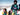 Two children in colorful snowsuits, harnesses, and sunglasses stand on a snowy mountain summit with alpine scenery in the background.