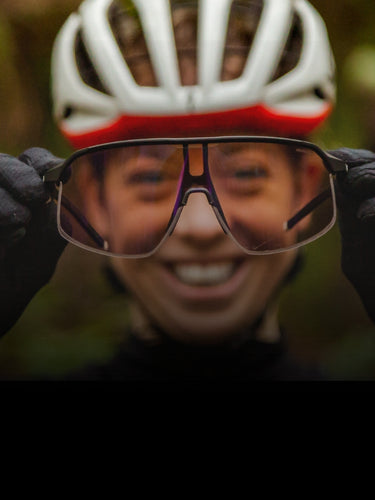 A person wearing a bike helmet and gloves holds up a pair of sports glasses in front of their smiling face, with the glasses in focus and the face blurred in the background.
