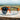A pair of sports sunglasses with orange and black frames is resting on a wooden surface outdoors.