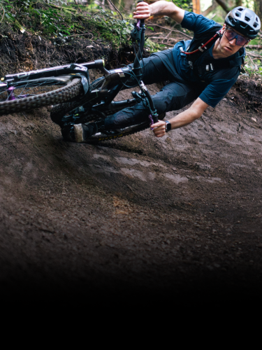 A mountain biker wearing a helmet and sunglasses leans sharply into a dirt trail turn in a forested area.