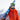 A skier in an orange jacket and helmet stands outdoors, smiling and holding a ski over his shoulder with snowy mountains in the background.