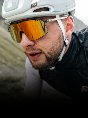 A man wearing a white bike helmet, reflective sunglasses, and a black cycling vest rides outdoors, with a blurred background of grassy hills.