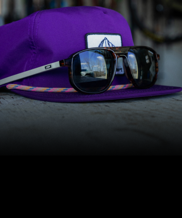 A pair of sunglasses resting on a purple baseball cap with a rectangular white patch, both placed on a dark surface.