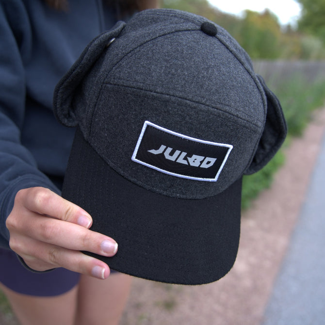 A person holding a dark gray and black cap with the word Julbo on a rectangular patch. The cap has ear flaps and is held toward the camera outdoors.