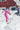 Two people in colorful snowsuits and sunglasses play in deep snow outside a red house, with one person snowboarding and the other raising an arm in the background.