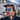 A bearded man in a black cap and fleece jacket wears Julbo Eyewear’s Legacy RX vintage-inspired sunglasses with blue reflective lenses, standing outdoors with mountains and buildings behind him.