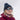 A smiling person wears Julbo Eyewear's Legacy RX round frames, paired with a maroon jacket, floral neck gaiter, and blue beanie, standing outdoors in a snowy landscape.