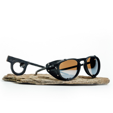 A pair of black Julbo Eyewear Limited Edition Millenium sunglasses with side shields and round arm cutouts, polarized Category 3 lenses, and a hand-forged piton bottle opener, displayed on a flat stone surface.