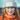 A person with red hair in braids wears a gray knit beanie and an orange jacket, smiling slightly while looking at the camera.