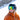 Person in an orange jacket and ski helmet with reflective goggles stands on a snowy mountain slope with rocky cliffs in the background.