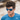 A man with tousled dark hair wears Julbo-usa Renegade blue adventure sunglasses with REACTIV photochromic lenses and a blue Adidas shirt, outdoors against a blurred background.