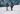 Two people in winter gear stand on skis on a snowy slope with a mountainous landscape and a village visible in the background.
