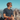 A man wearing Julbo-usa Shield eyewear and a t-shirt sits on a rocky mountain with distant peaks in the background under a clear sky.