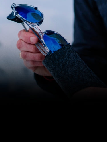 A person holds a pair of blue-lensed sunglasses, partially removed from a dark felt case.