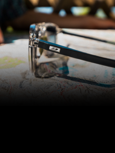 Close-up of transparent glasses with black arms resting on a map, with blurred background featuring a wooden fence and out-of-focus person.