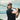 A man wearing Julbo Eyewear's The Streets RX sunglasses, a cap, and a black shirt holds a golf club over his shoulder outdoors by water under a cloudy sky.