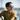 A person with short dark hair wears Julbo-usa Vermont Classic sunglasses and a yellow shirt, looking to the side outdoors on a sunny day with blurred water and sky in the background.