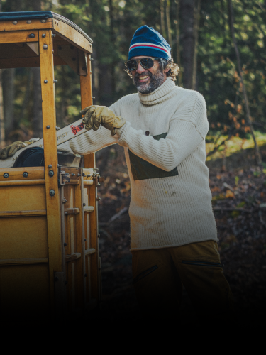 A man in a white sweater, yellow pants, and blue hat stands outdoors by a yellow vehicle, smiling while holding a handsaw. Trees and foliage are visible in the background.