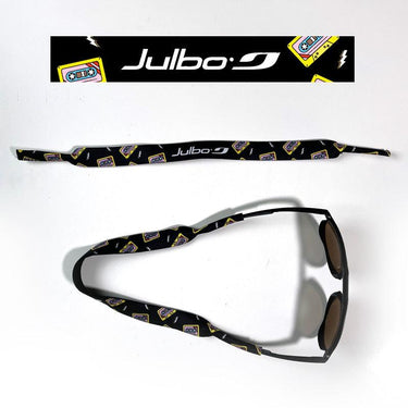 The Julbo Eyewear Neoprene Sunglass Strap is black with yellow cassette tape graphics, offers a waterproof, universal fit for secure comfort, and retro style. Shown both laid flat and attached to sunglasses. #color_julbo-neoprene-sunglass-strap-black-cassette-tapes