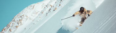 A skier in tan gear and black helmet skis downhill on a steep, snowy mountain slope, sending up a spray of powder.