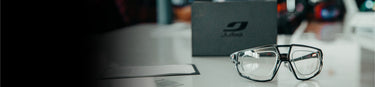 A pair of eyeglasses is placed on a white surface next to a gray Julbo box and a sheet of paper.