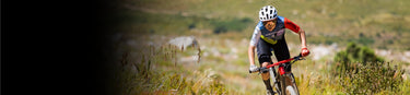 A cyclist wearing a helmet and sports gear rides a mountain bike on a grassy outdoor trail with blurred greenery in the background.