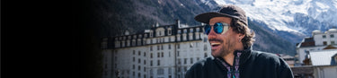 A man with sunglasses and a cap smiles outdoors with a snowy mountain and buildings in the background.