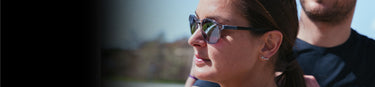 A close-up of a woman wearing sunglasses and earrings outdoors, with part of another person visible behind her.