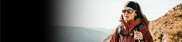 A person wearing sunglasses and outdoor gear hikes on a mountain trail under a clear sky, with hills in the background.