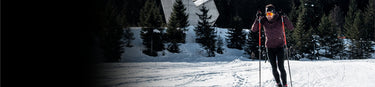 A person wearing winter gear and sunglasses cross-country skiing on a snowy trail with pine trees and a wooden structure in the background.
