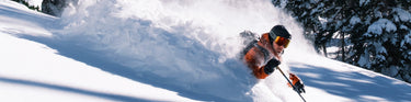 A skier in orange gear moves quickly through deep powder snow on a sunny, tree-lined slope.