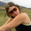 A woman outdoors in a grassy field with mountains in the background, wearing black-framed sunglasses with orange-tinted lenses and a black tank top.
