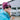 A smiling woman wearing a burgundy Teton beanie, black sunglasses, and a bright turquoise jacket, photographed outdoors with a blurred building in the background.
