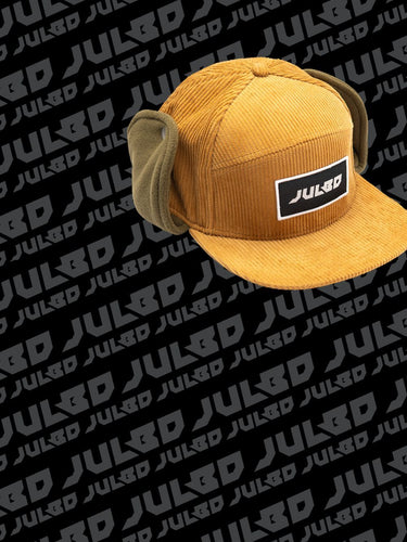 A mustard yellow corduroy cap with ear flaps and a JULBO logo patch, placed on a background patterned with repeating JULBO text.