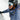 A close-up action shot of a person in a snowy outdoor environment wearing a blue puffer jacket, black helmet, mirrored ski goggles, and an orange neck gaiter.