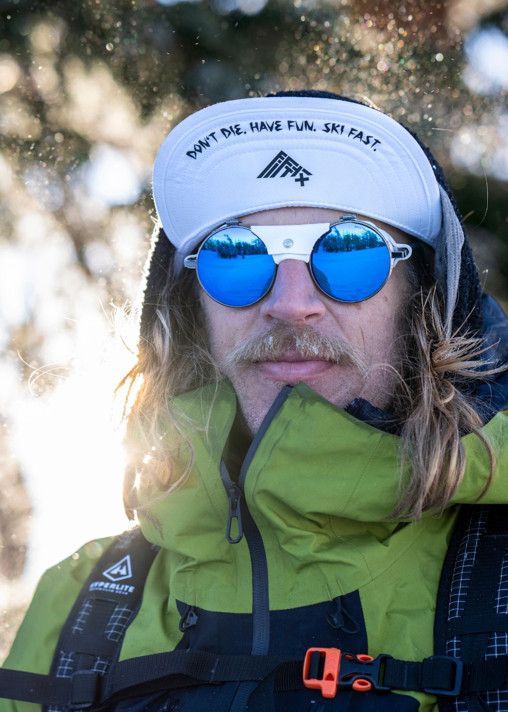 Person wearing a green jacket, white hat with the text DONT DIE. HAVE FUN. SKI FAST., and mirrored sunglasses, standing outdoors in a snowy setting.