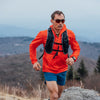 Man running on a mountain trail wearing an orange jacket and sunglasses.