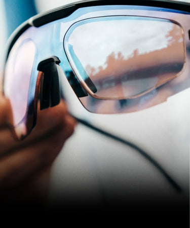 Close-up of a hand holding a pair of sporty sunglasses with mirrored lenses reflecting an outdoor scene.