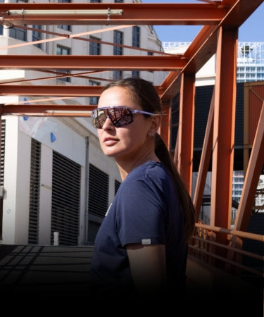 A woman wearing sunglasses and a navy shirt stands outdoors under a red metal structure with buildings in the background.