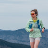 Woman in a light green jacket and sunglasses running with mountains in the background