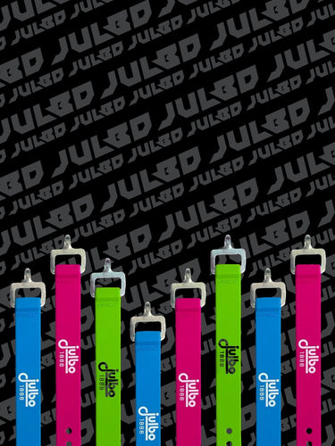 Six colorful Julbo ski goggle straps—three pink, two green, one blue—stand upright in front of a repeating JUL&BD patterned black background.