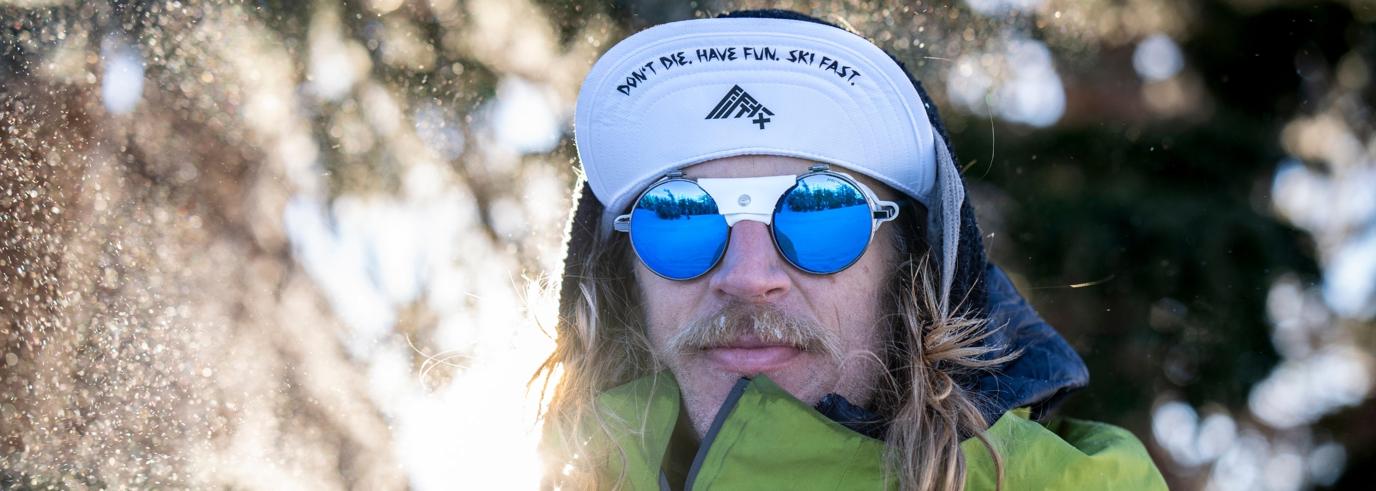 A person wearing reflective blue sunglasses, a white hat reading DONT DIE. HAVE FUN. SKI FAST., and a green jacket stands outdoors in snowy, sunny conditions.