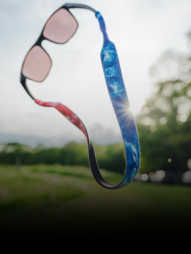 A pair of sunglasses with a colorful blue and red strap appears to float outdoors, with trees and greenery blurred in the background.
