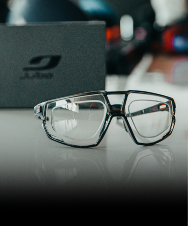 A pair of black-rimmed eyeglasses is displayed on a white surface with a branded box and blurred background objects behind them.
