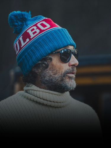 A man with curly hair and a beard wears sunglasses, a blue and red knit hat, and a cream turtleneck sweater, standing outdoors.