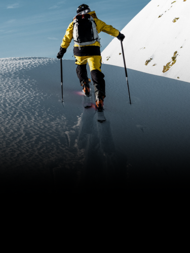 Person wearing yellow jacket and backpack hiking uphill on snow-covered mountain with ski poles under a clear blue sky.