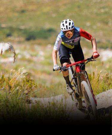A cyclist wearing a helmet and cycling gear rides a mountain bike uphill on a dirt trail with grassy terrain in the background.